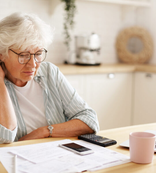 People, age, technology and finances. Depressed unhappy retired woman paying domestic bills online, trying hard to make both ends meet, sitting at kitchen table, surrounded with papers, using gadgets